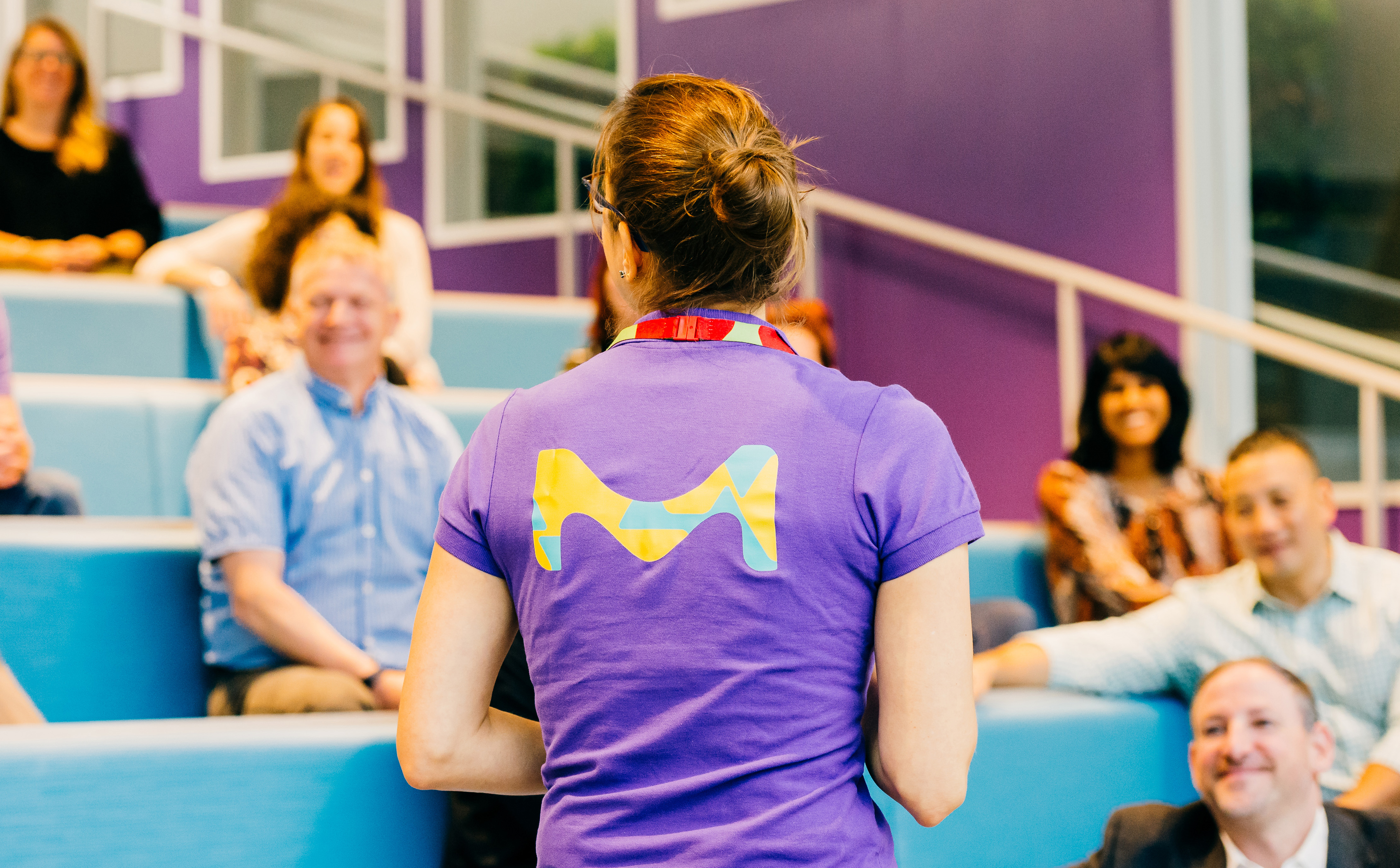 The Future of Organoids Is Streaming! A person with brown hair in a bun, wearing a purple T-shirt with a colorful, abstract “M” logo on the back, stands facing an audience in a blue-seated auditorium with purple walls.