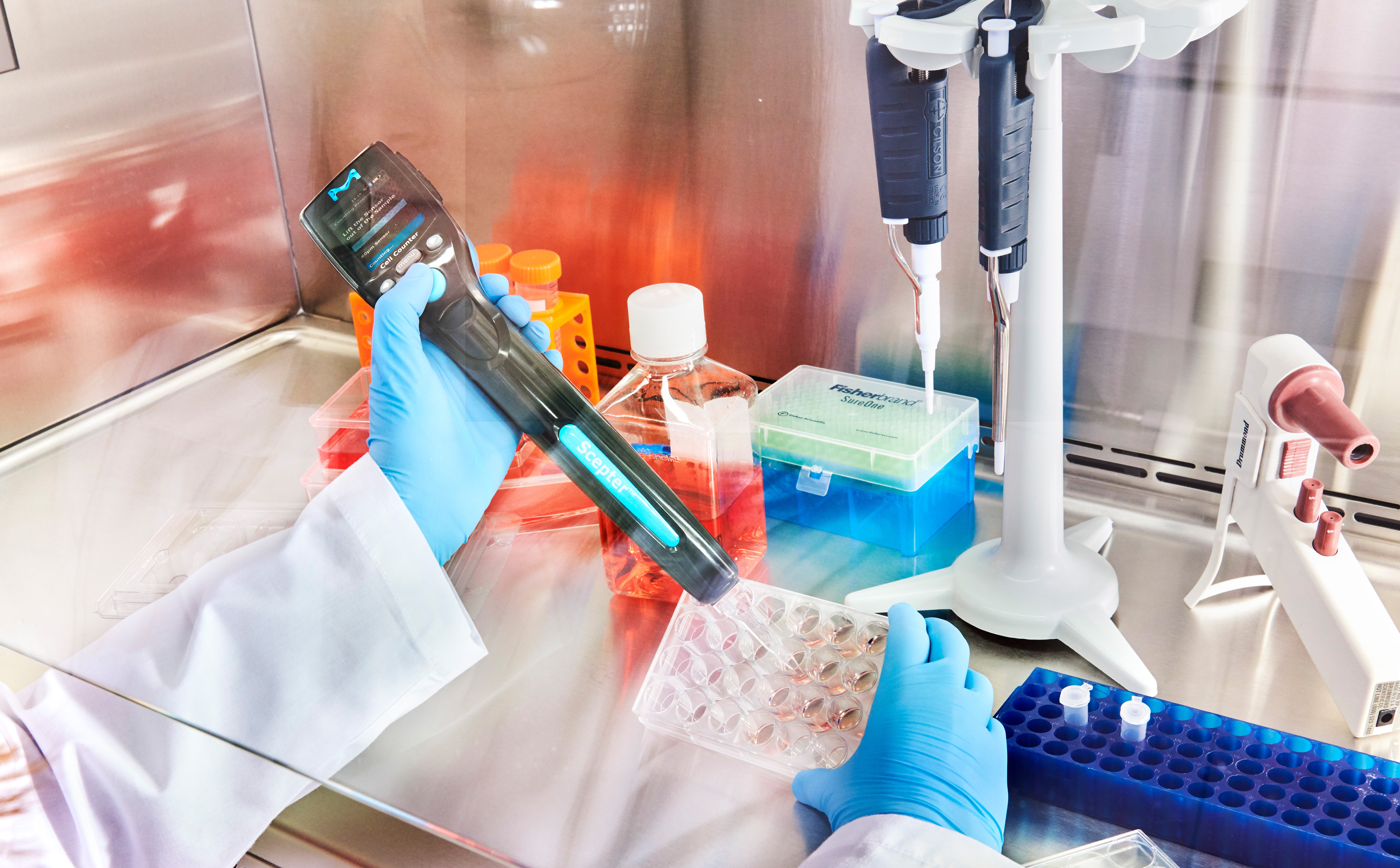 Win a Scepter Cell Counter! A scientist wearing blue gloves and a white lab coat is handling a handheld device inside a biosafety-style lab bench. They are near a rack of test tubes, a blue-tipped pipette stand, and other lab equipment, including a pipette and reagent bottles.