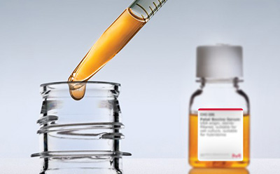 Serum bottle in the background halfway filled with golden liquid. In the foreground, a pipette partially filled with gold liquid is pulling out of a bottle top and dripping. Serum bottle in the background halfway filled with golden liquid. In the foreground, a pipette partially filled with gold liquid is pulling out of a bottle top and dripping.