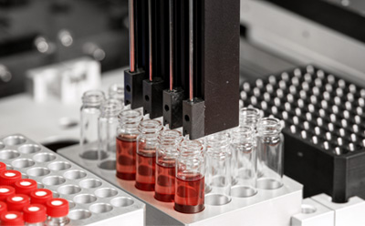 Vista en primer plano de un sistema de automatización de laboratorio con múltiples puntas de pipeta colocadas sobre varios viales de vidrio que contienen líquido rojo.