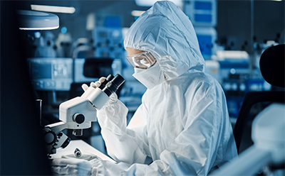 Pyrogen Testing A researcher in a full protective suit and goggles examines a sample under a microscope in a laboratory setting, with various scientific equipment visible in the background.