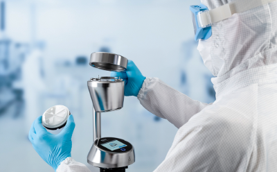 Viable Air Monitoring New A scientist in a protective suit and gloves examines a petri dish containing a sample, holding it up to the light against a blurred background of laboratory equipment.