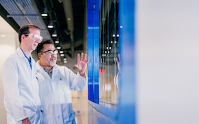 Two laboratory technicians in white lab coats are engaged in a discussion while looking at a large display screen. One technician is gesturing with their hand, indicating a point of interest, while the other is attentively listening.