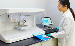 Figure 1 A laboratory benchtop bioprinting system is shown inside a clean research environment. The enclosed instrument features multiple printheads positioned above a printing stage within a transparent chamber. A researcher in a laboratory coat is seated beside the instrument, operating the system through a connected computer workstation.
