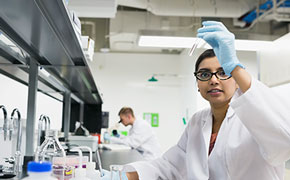 In a modern laboratory with bright white walls and ceilings, a female scientist wearing a white lab coat, blue latex gloves, and black-framed glasses is carefully examining a clear glass test tube filled with a transparent liquid. She holds the test tube up to eye level with a focused expression, ensuring precise observation. The workstation in front of her contains various laboratory glassware, including flasks and beakers filled with solutions, as well as plastic containers with red and yellow lids. Stainless steel faucets and a black lab bench are visible, emphasizing a well-equipped analytical environment. In the background, a male scientist in a white lab coat is working at another station, slightly out of focus, contributing to the dynamic and professional atmosphere of the laboratory.