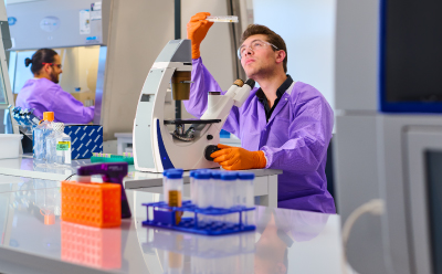 Lab technician in a purple coat and orange gloves using a microscope at a biosafety testing bench, with tubes and lab equipment visible in the background.