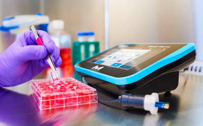 ERS 3.0 in the tissue culture hood Scientist’s hand holding tweezers picking up a clear insert for a cell culture plate filled with culture media in the tissue culture hood. ERS 3.0 instrument is directly to the right and displays the culture plate map. ERS 3.0 probe is laying directly in front of the instrument.