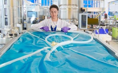 Ultimus® film A person in a lab coat and purple gloves stands behind a large, transparent blue plastic container filled with liquid. Tubing is connected to the container, and the background shows various laboratory equipment and machinery.