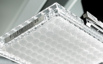 A close-up view of a MultiScreen<sup>®</sup> 96-well plate with a barcode label, held by a robotic arm, showcasing the circular wells used for sample analysis.