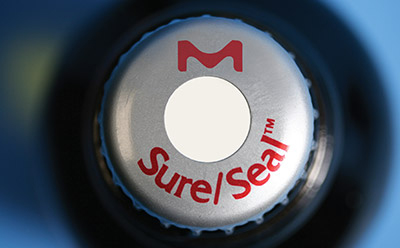 Close-up of a bottle cap featuring a logo with an ‘M’ above a white circle, and the text “Sure|Seal™” in red around the lower edge of the circle. 