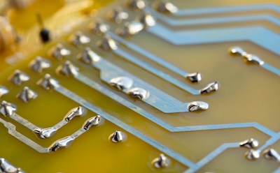 High Purity Metals in Electronics and Semiconductors 一张电子电路板的特写照片,板上布满了导线。这些导线在鲜艳的黄色背景衬托下,因闪亮的银色焊锡滴而格外醒目。