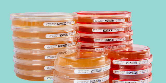 <p>¿Qué importancia tiene la selección de los medios de cultivo en la vigilancia medioambiental?</p> Pilas de placas de cultivo etiquetadas de color naranja y rojo dispuestas sobre un fondo turquesa.