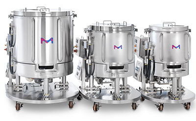 Three stainless steel bioreactor vessels of varying sizes on wheeled stands, each with a blue and pink ‘M’ logo on the front.