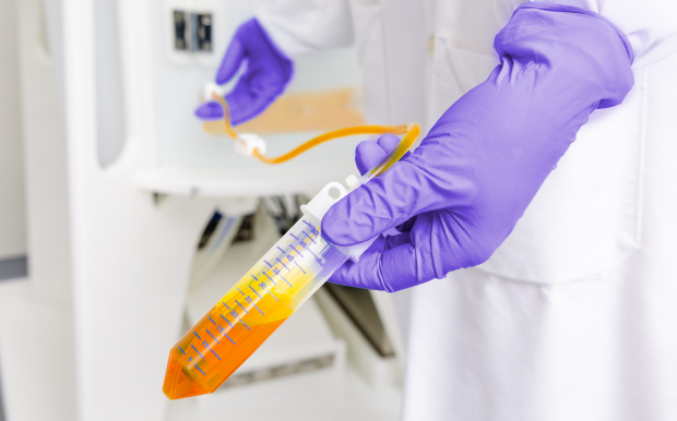 A laboratory worker wearing purple gloves holds a NovaSeptum® SURe Sampling Assembly with transparent tubing and orange fluid in a single-use processing environment
