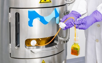 A lab technician wearing a white coat and purple gloves operates a NovaSeptum® SURe sterile sampling assembly connected to a stainless-steel bioprocess vessel, with a sample container hanging from the tubing.