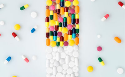 The image depicts an array of colorful pills and capsules artistically arranged on a white surface to resemble the shape of a pill bottle, with the body composed of multi-colored capsules and the cap made of white round tablets, surrounded by additional loose pills.