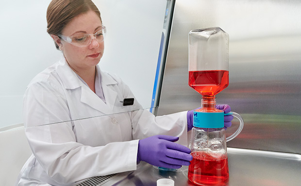 The image show a woman in a white lab coat, working with laboratory flask containing a red liquid, which is connected to another piece of equipment with blue liquid at the bottom.