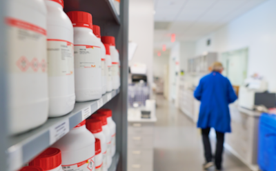 Sigma-Aldrich white bottles with a red cap are on the right side. A researcher walks in a lab away from the bottles on the right.