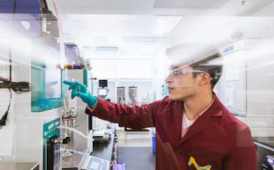 Scientist wearing a lab coat with a vibrant M, gloves, and safety glasses reaches towards an instrument and adjusts settings.