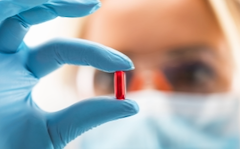 A red pill is in the foreground. In the background, a blurry image of someone holding the pill wearing a mask and gloves