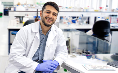 Scientist in a lab environment wearing a lab coat and protective gloves sitting at desk 