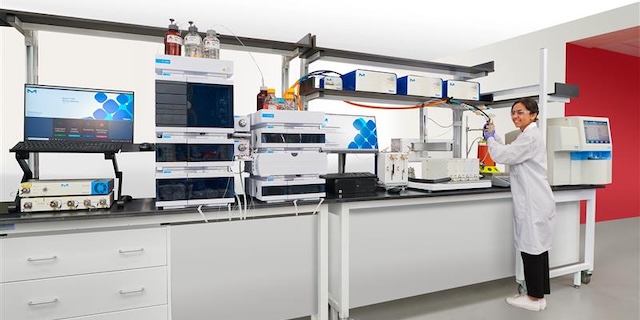 A laboratory setting featuring advanced scientific equipment, including multiple chromatography systems and a computer monitor displaying data. A researcher in a lab coat is actively engaged in an experiment at a workbench, surrounded by various instruments and supplies essential for laboratory analysis.