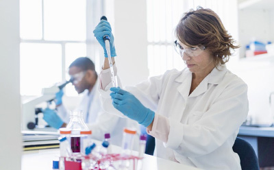 Scientist pipetting a sample at a lab bench while another scientist uses a microscope in the background.