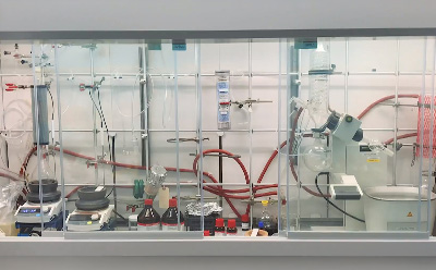 A laboratory fume hood displaying an organized organic synthesis workstation, including hot plates, condensers, glassware, tubing, reagent bottles, and a rotary evaporator arranged for conducting and optimizing chemical reactions.