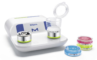 Milliflex Oasis® Filtration System The Millipore Milliflex Oasis® filtration system, featuring a sleek white base with two circular filtration units and a transparent container on top, accompanied by several Petri dishes in blue and pink, all arranged on a clean, white background.