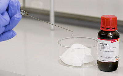 A gloved hand holds a syringe releasing a small droplet above a clear glass dish, with a labelled amber bottle of organolithium solution placed nearby on the laboratory benchtop.