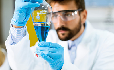 A laboratory researcher wearing protective gloves carefully operates a separatory funnel containing a blue liquid, with additional lab equipment visible in the background.