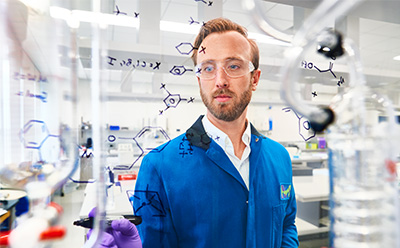 A laboratory scientist in a blue lab coat writes chemical structures on a transparent glass surface, surrounded by lab equipment and instrumentation in a bright, well organized research space.
