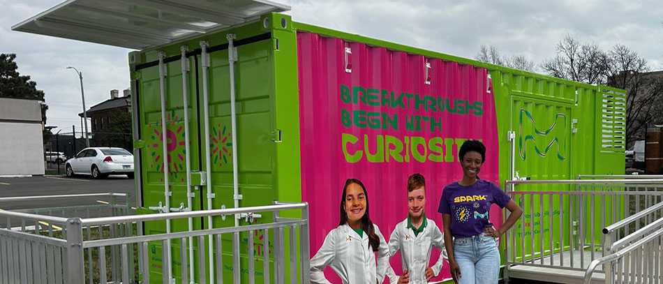 Danielle Chavis in front of the Curiosity Cube Danielle Chavis wears a SPARK t-shirt and stands in front of the Curiosity Cube which is bright green, has images of two young scientists with the words “Breakthroughs Begin with Curiosity” on the front.