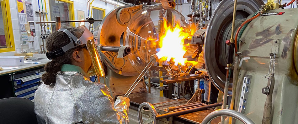 Glassblower works on flask 丸底フラスコは旋盤に固定され、ガラス器具職人がフラスコのネック部に直接炎をあてている。ガラス器具職人は特殊なジャケット、グローブ、フェイスシールドなどの防護服を着用している。