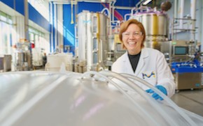 Shannon Cleveland Holds Ultimus Bag Shannon wears a lab coat, blue gloves and safety glasses and stands in an M Lab — a non-GMP biomanufacturing setting. She is smiling and holding onto a bin bag, which is situated in the lower left corner.