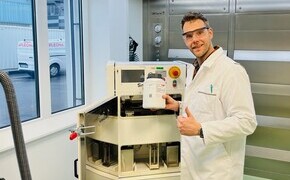 Thomas Kipping in lab - tile Thomas Kipping wears safety glasses and a lab coat standing next to a laser 3D printer.