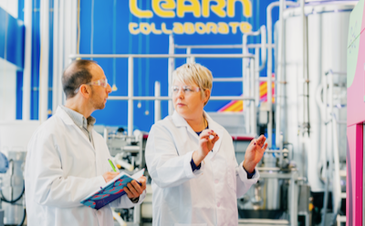 Two researchers wear safety glasses and stand next to large bioreactors