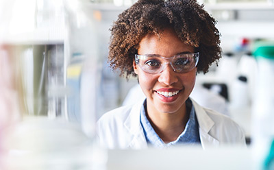 A person wearing a lab coat and safety glasses is working in a laboratory setting, surrounded by scientific equipment and glassware. The background features blurred laboratory elements, emphasizing a professional research environment.