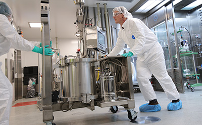 MP cleanroom expertise Two GMP-gowned technicians in white Tyvek suits transporting a stainless steel bioreactor in a pharmaceutical cleanroom for difficult-to-purify small-molecule compound processing