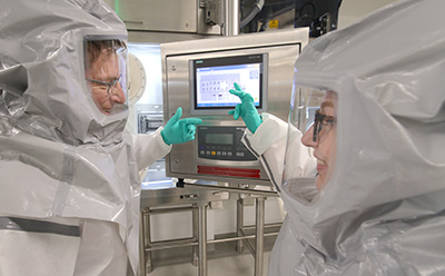 Small molecule development process Two pharmaceutical scientists in grey PAPR suits reviewing a Siemens SIMATIC HMI control interface, illustrating the collaborative CDMO engagement process for small-molecule projects