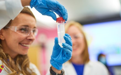 Innovative strategies for nitrosamine prevention Close-up of a laboratory professional wearing safety glasses and blue gloves holding a sample tube containing liquid while another colleague stands in the background.