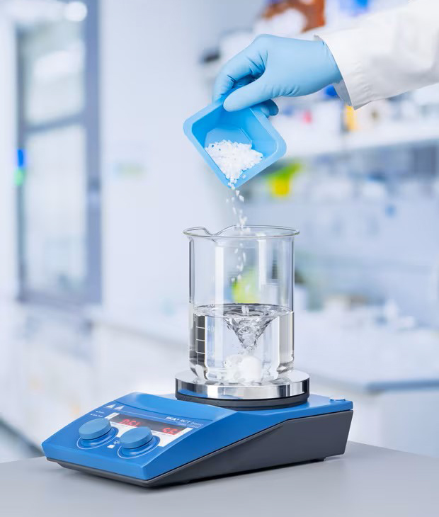 Gloved hand transferring a white powder sample into a beaker placed on a digital analytical balance in a laboratory setting.