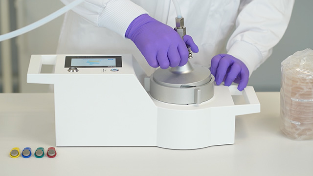 <p>Muestreador de gases comprimidos microbianos MAS-100 Atmos®</p> Primer plano de unas manos con guantes morados que manejan un muestreador microbiano de gas comprimido en un instrumento de laboratorio.