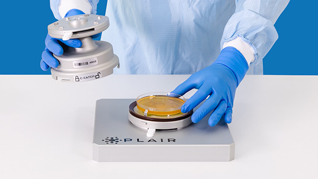 <p>Sistema de control de la calidad del aire Rapid-C+</p> Unas manos con guantes azules colocan una placa de cultivo en un dispositivo de control de la calidad del aire P L A I R, mientras sujetan su componente de muestreo.