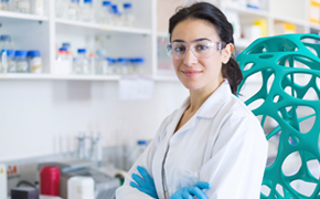 A person in a white lab coat and blue gloves stands confidently with arms crossed in a laboratory setting. Behind them are shelves filled with various laboratory supplies and equipment, indicating a professional research environment.