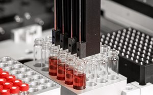 A close-up view of a laboratory automation system featuring multiple robotic arms positioned above several glass vials containing red liquid. The vials are arranged on a tray with circular openings, and the background includes various laboratory components and equipment.