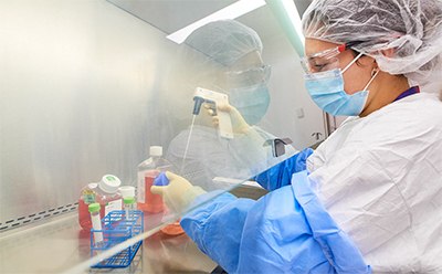Pathogen and Spoilage Testing Webpage Picture A person in protective clothing, including a hairnet and face mask, working in a laboratory with various bottles of chemicals on the countertop. Background with a clean, organized workspace, including a laptop and scientific equipment.