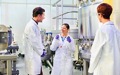Tres individuos en batas de laboratorio en un entorno de laboratorio bien equipado. La persona que está en el centro está tiene una conversación con el individuo de la izquierda. La persona de la derecha está observando. Tres individuos en batas de laboratorio en un entorno de laboratorio bien equipado. La persona que está en el centro está tiene una conversación con el individuo de la izquierda. La persona de la derecha está observando.