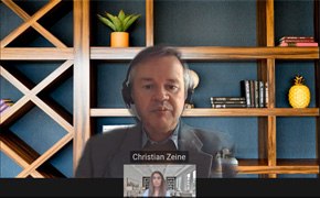 Una captura de pantalla de una reunión virtual con Christian Zane y otro participante. Una pequeña ventana muestra al otro participante hablando. Al fondo, un estante bien organizado contiene libros, objetos decorativos y una maceta.
