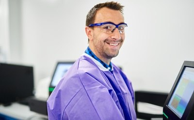 A laboratory technician wearing a purple lab coat is seated at a workstation with computer monitors displaying data. The background features additional computer setups.